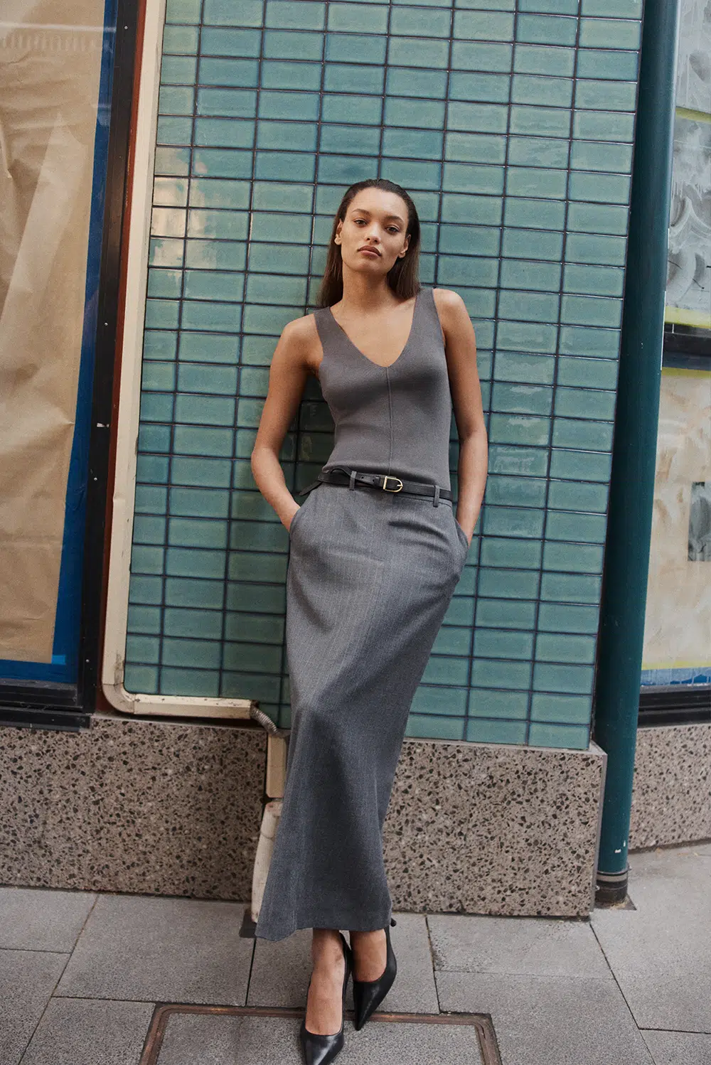 Elegant woman in grey dress and black heels standing against a blue tiled wall, fashion pose, outdoor setting, natural light.