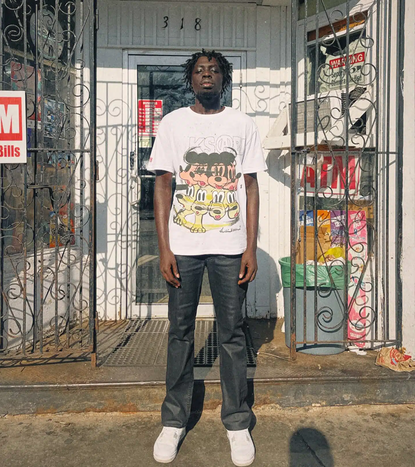 An adult male stands in front of a building wearing a cartoon graphic t-shirt, jeans, and sneakers outside during the daytime.