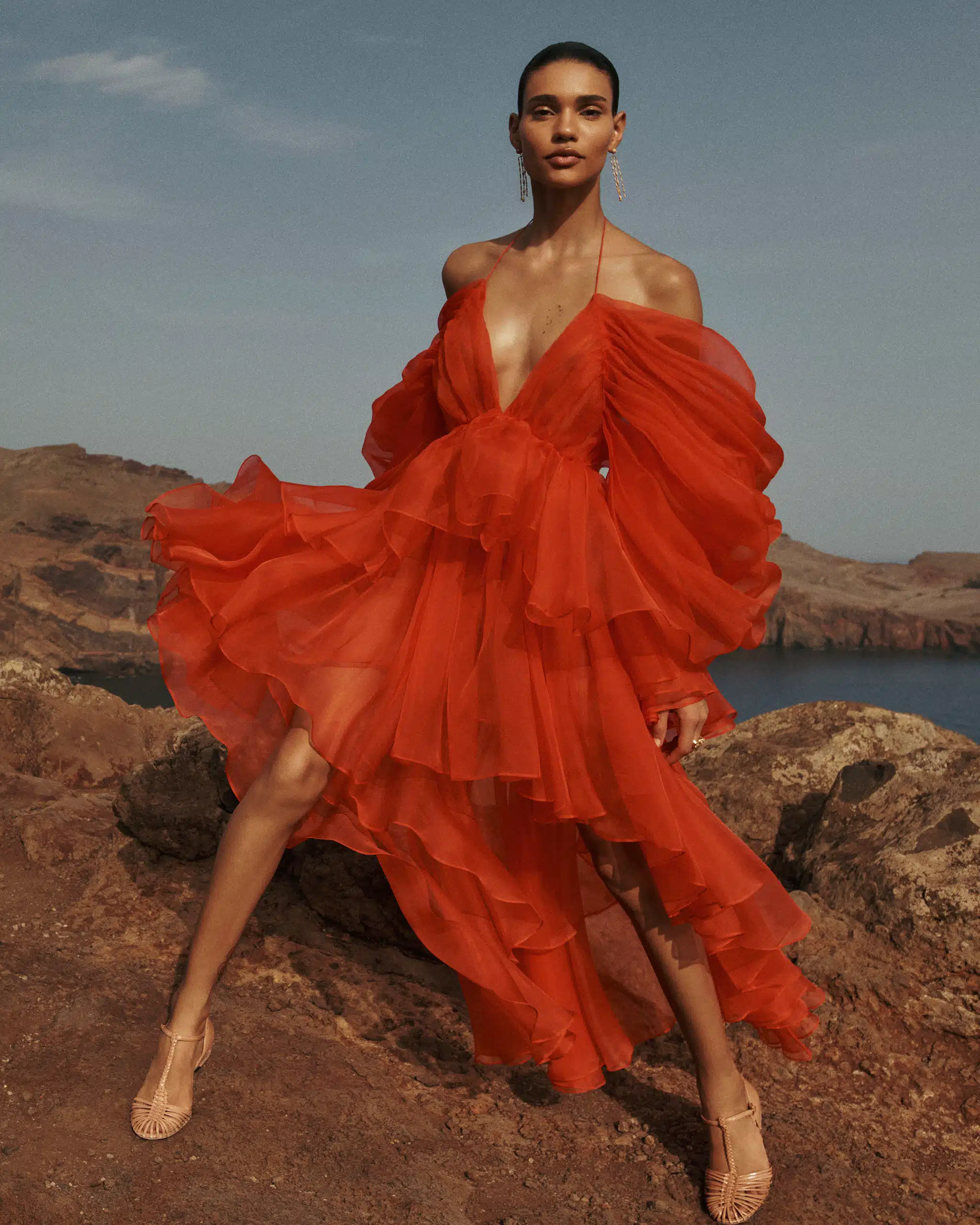 A woman in a red, tiered dress stands on a rocky landscape near the sea, under a blue sky, in a fashion editorial style photograph.