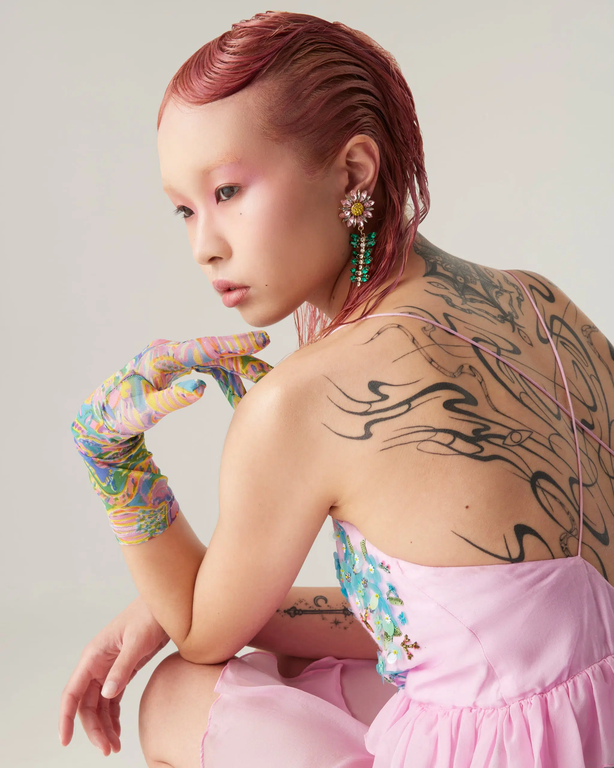 Asian woman with red hair, side view, wearing a pink dress, colorful gloves, and jewelry poses in a photo studio against a neutral backdrop.