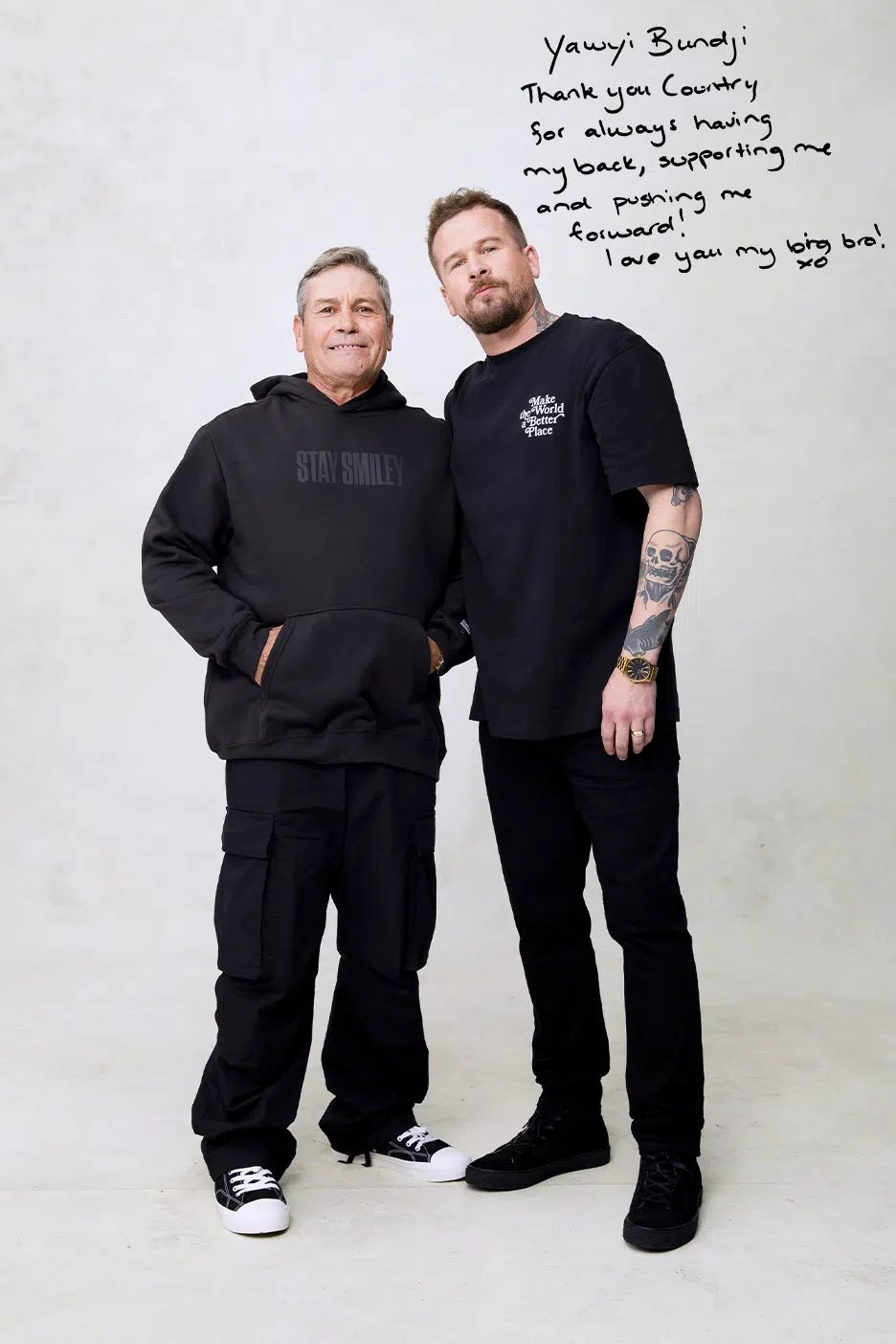 Two men stand side-by-side in black casual attire against a white background, with handwritten text visible in the upper right corner.
