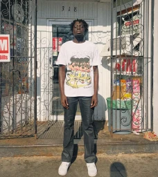 A Black man standing in front of a storefront wearing a white graphic t-shirt and dark trousers, captured by Emil Vrisakis.