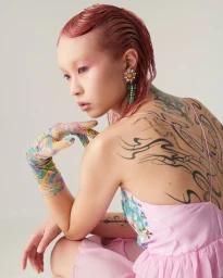 Model with pink hair and back tattoos wearing a light pink dress and patterned sheer gloves against a plain white background.