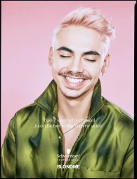 Portrait of a man with pink hair and a mustache smiling, wearing an iridescent green satin shirt against a pink backdrop