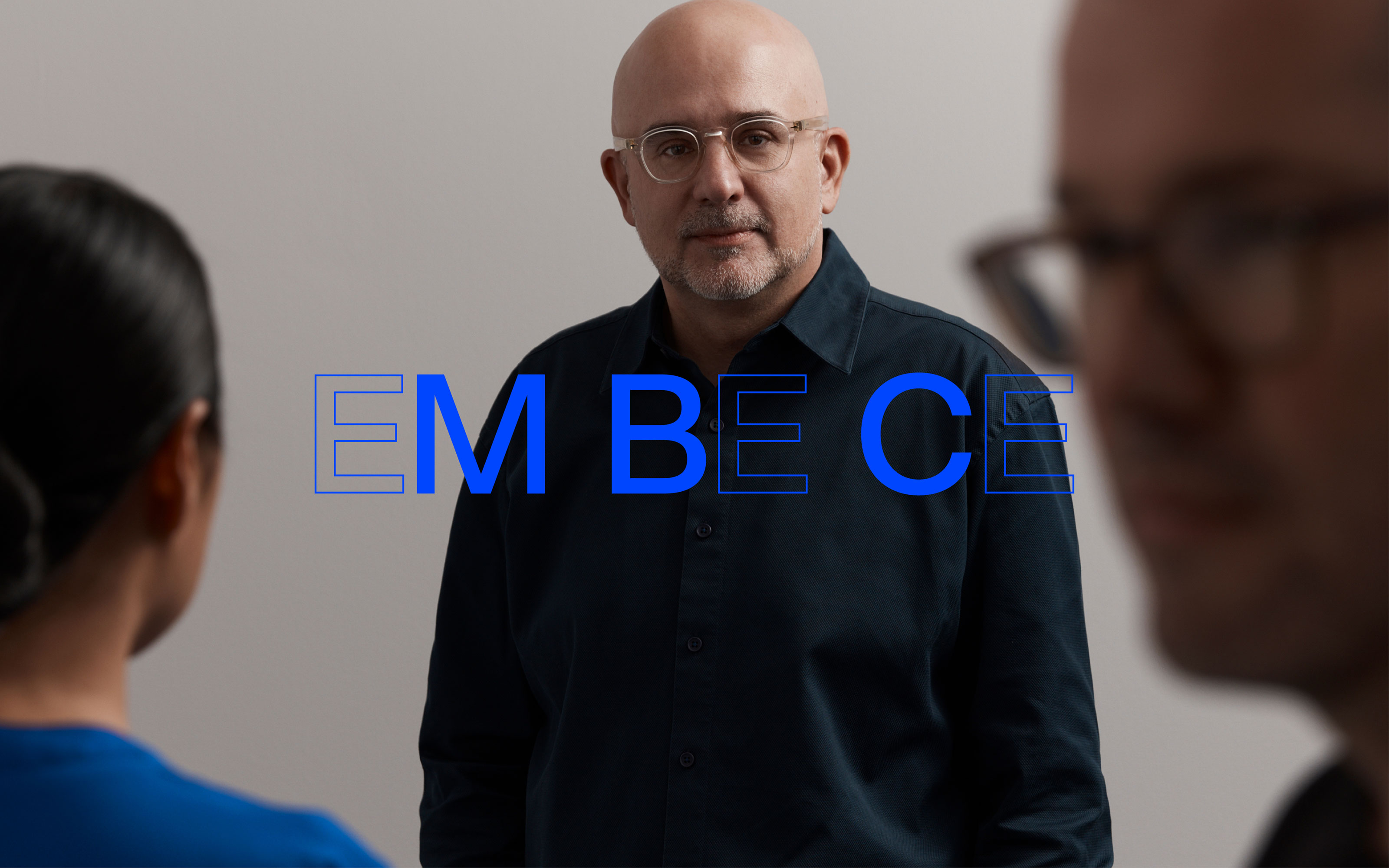 Indoor shot of two adults looking toward a bald adult male with glasses in a button-down shirt against a light background.