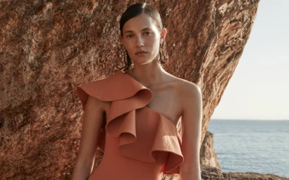 Model in rust-coloured one-shoulder ruffled top standing in front of rocky coast with ocean background
