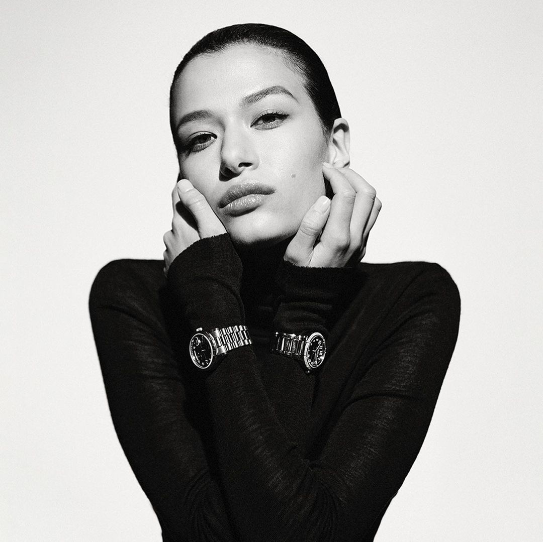 Monochrome portrait of a woman wearing a black turtleneck and two wristwatches, hands near her face, against a bright background.