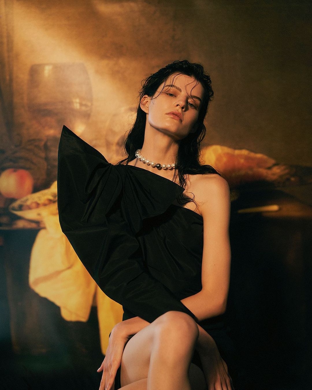 A woman poses in a dark, draped dress with a pearl necklace against a backdrop reminiscent of a classic still-life painting.