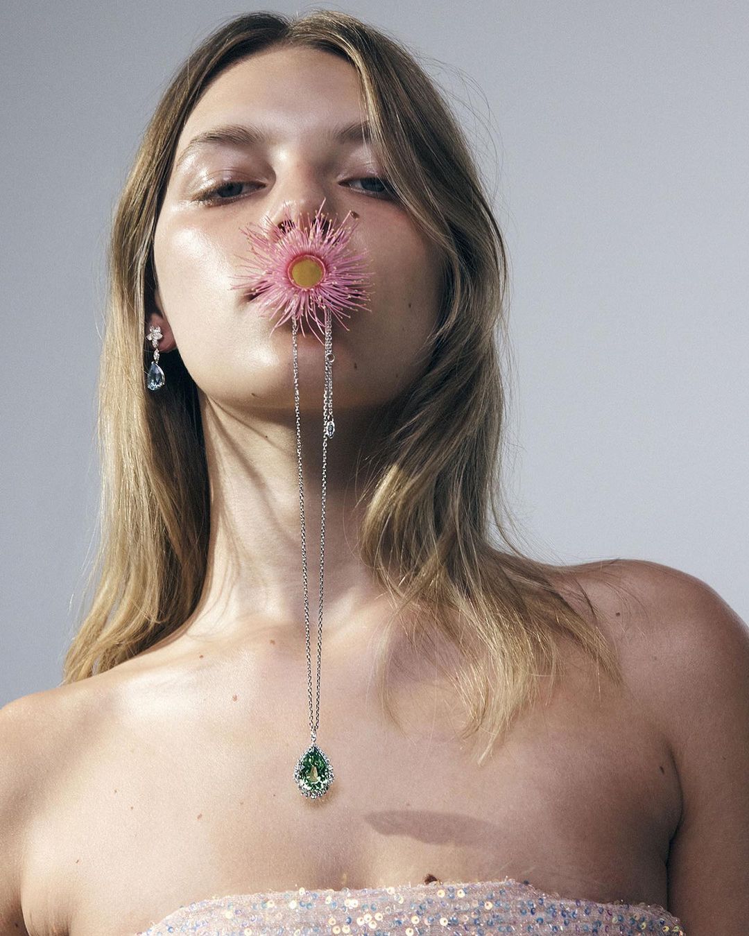 Close-up of a blonde woman with light skin, wearing a pink strapless dress, posing with a flower in her mouth and green pendant necklace.