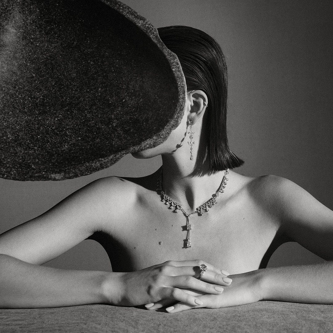 Monochrome fashion shot of a woman wearing jewelry, with a large rock partially covering her face, in a neutral indoor setting.