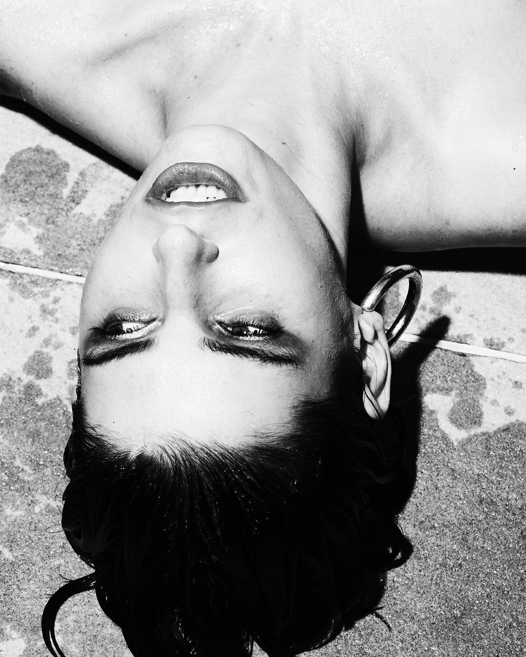 Black-and-white portrait of a dark-haired woman lying on her back with large, round earrings and a neutral expression on a gray surface.
