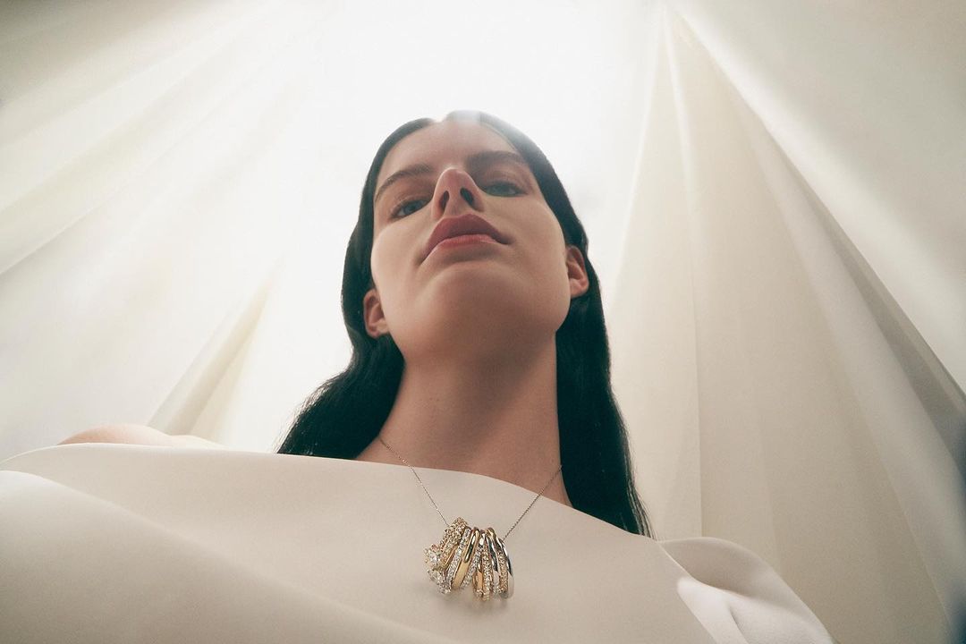 Low-angle shot of a woman with black hair wearing a white top and statement jewelry against a soft, white background.
