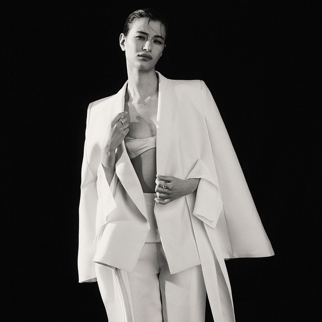 Monochrome fashion portrait of a slender model in white pantsuit and bikini top posing in front of a dark background.