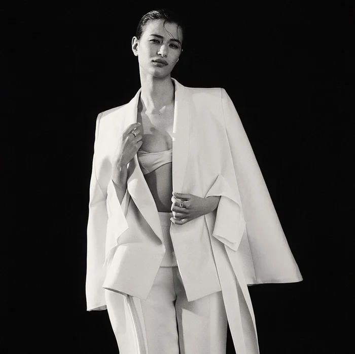 Monochrome portrait of a woman in a white suit jacket worn draped over her shoulders against a black background.