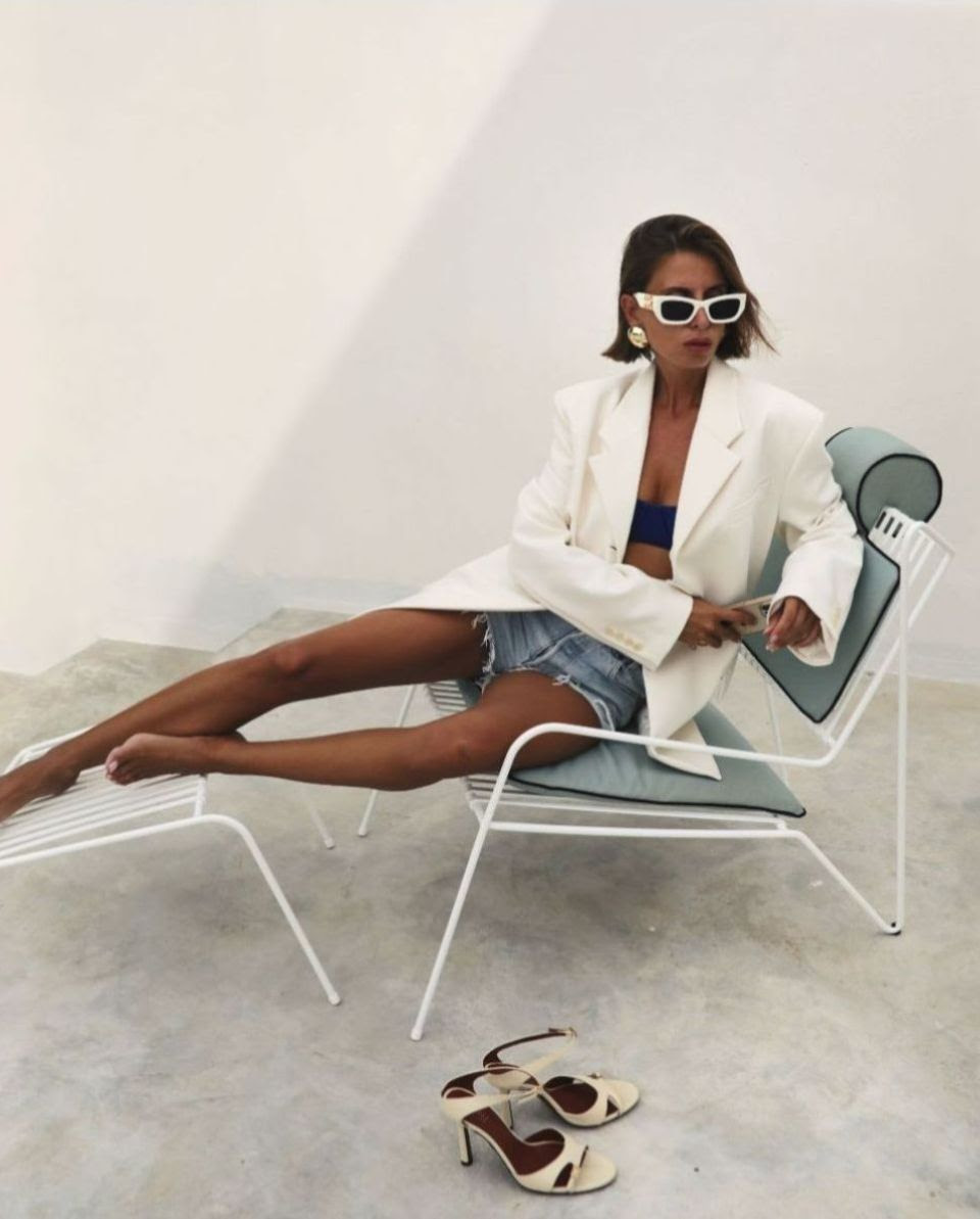 Woman in sunglasses and white blazer relaxes on metal lounge chair with sandals on floor against white wall in fashion shot.