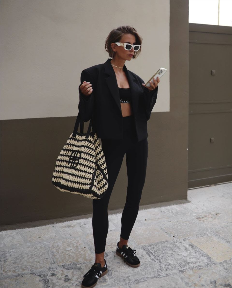 Woman in black blazer and leggings holds phone and wears sunglasses, standing against a beige wall with a striped tote bag.