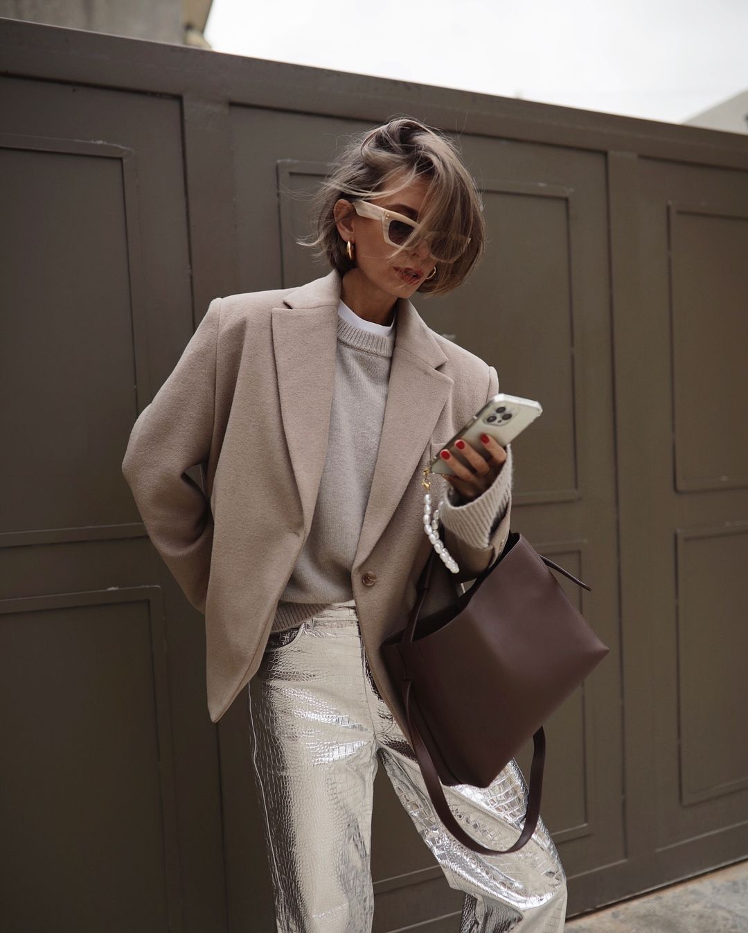 A stylish adult woman in a beige blazer and sequin pants uses a smartphone and carries a brown purse in an outdoor urban setting.
