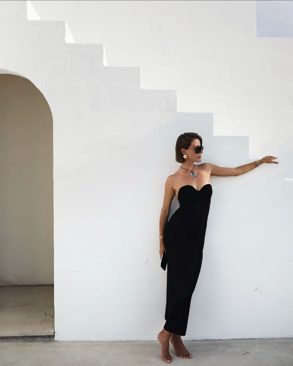 Woman in black strapless dress poses on outdoor white staircase in sunglasses and jewelry, creating a modern, elegant fashion portrait.