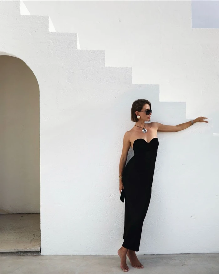 Woman in a black strapless midi dress leaning against a white architectural wall with stepped features