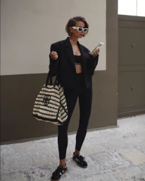 Woman in black blazer and leggings holding phone outdoors, carrying a woven black and white tote bag.