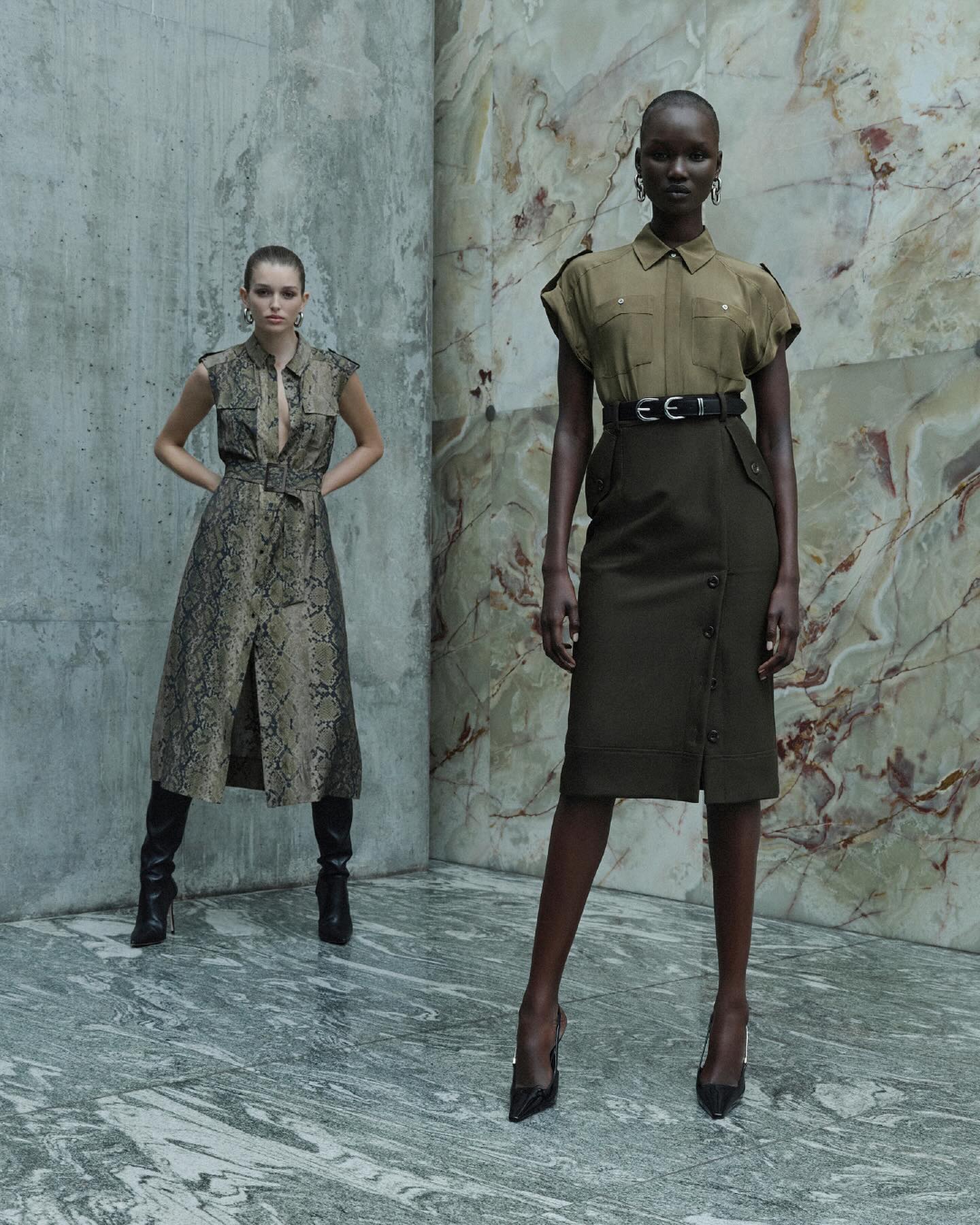 Two women pose indoors against mottled marble and concrete walls. One woman is Black in a utilitarian-style midi skirt and button-down shirt.
