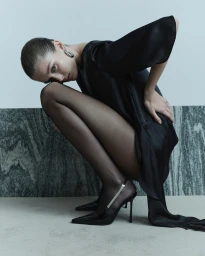 Model in black dress and sheer tights squatting against a marble wall for Cue SS24