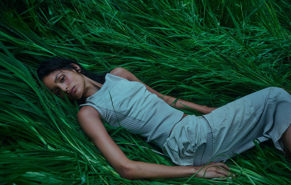 Adult woman lying in tall green grass wearing light gray top and skirt, eyes closed, natural light, relaxed pose, outdoor setting.