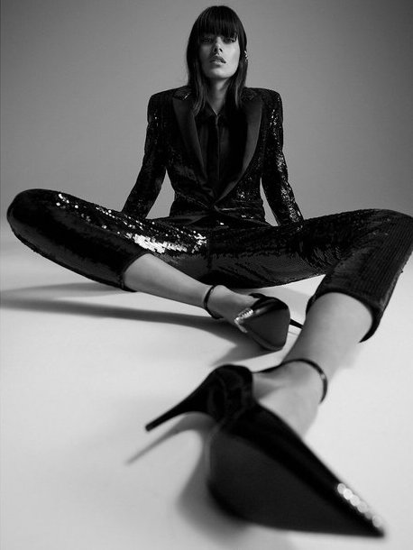 A woman in a sequined suit and heels poses in a studio with her legs spread in a black-and-white fashion shot, highlighting her stylish outfit.
