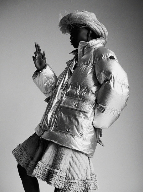 Black-and-white portrait of a Black woman in a metallic silver puffer jacket and skirt, accessorized with a feathered hat, against a plain background.