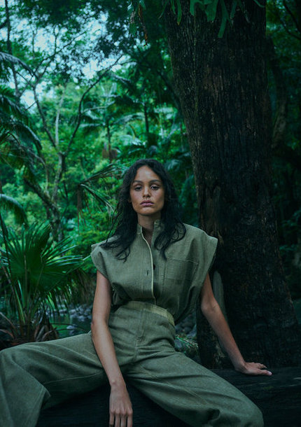 Adult female in olive jumpsuit sits on log in tropical forest; green tones, medium shot.