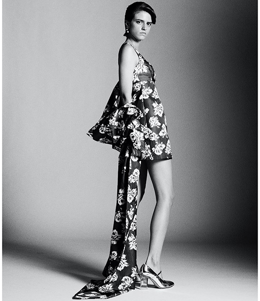 Monochrome fashion portrait of a woman in a floral dress and silver shoes in a studio with a neutral gray background.