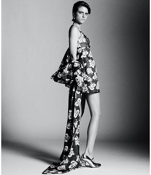 Monochrome fashion portrait of a woman in a floral dress and silver shoes in a studio with a neutral gray background.