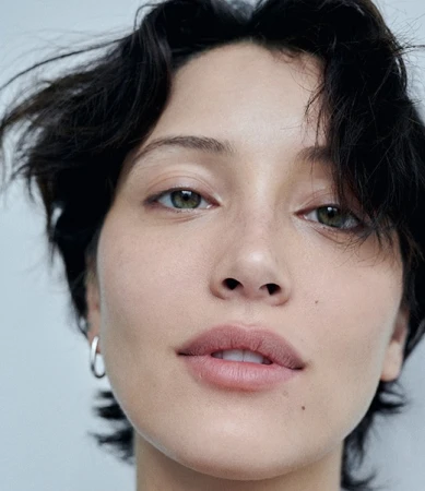 Close up portrait of a woman with short dark hair and green eyes wearing a silver hoop earring against a light background