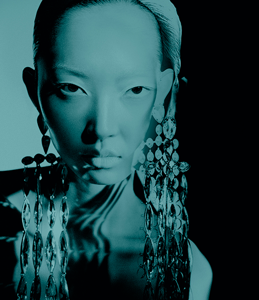 Close-up portrait of an Asian woman with pale skin wearing large crystal earrings against a black and turquoise gradient background.
