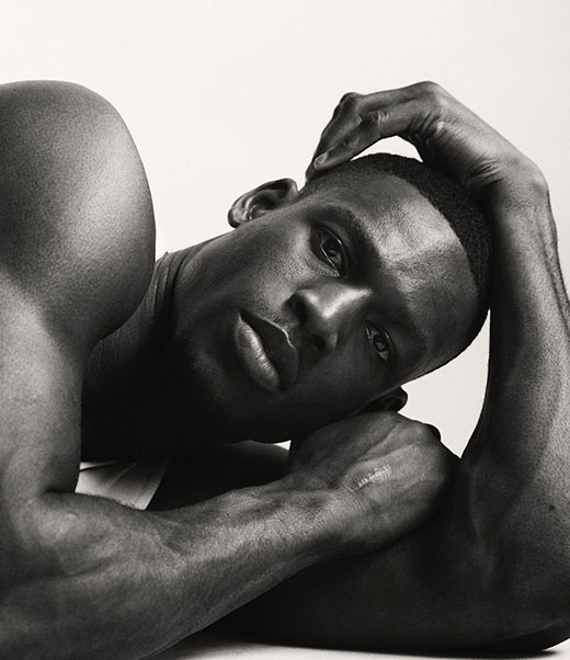 Black and white portrait of a muscular African-American man reclining, resting a hand on his head against a clean, bright background.