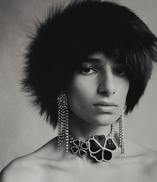 Portrait of a person wearing a dark fur hat and a flower-shaped diamond necklace and earrings in a photo studio setting.
