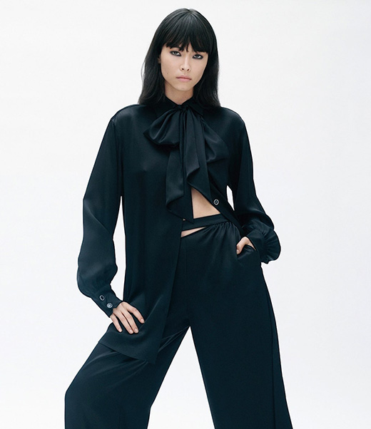 Fashion model in black silk blouse and wide leg pants poses in a photo studio against a white backdrop in a medium shot.