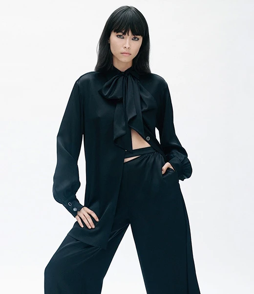 Fashion model in black silk blouse and wide leg pants poses in a photo studio against a white backdrop in a medium shot.
