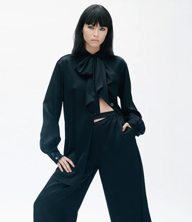 Fashion model in black silk blouse and wide leg pants poses in a photo studio against a white backdrop in a medium shot.