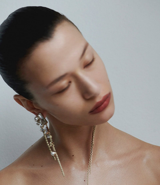 Close-up shot of a female fashion model with closed eyes, wearing large decorative earrings and a necklace, against a neutral backdrop.