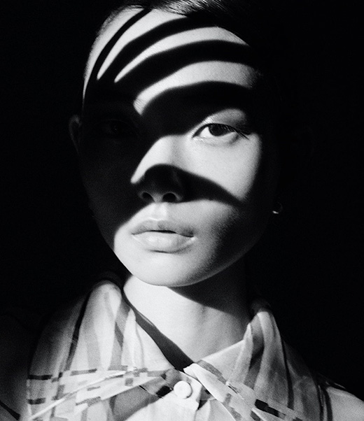 Monochrome portrait of an Asian woman with striped shadows across her face, wearing a striped blouse against a dark background.