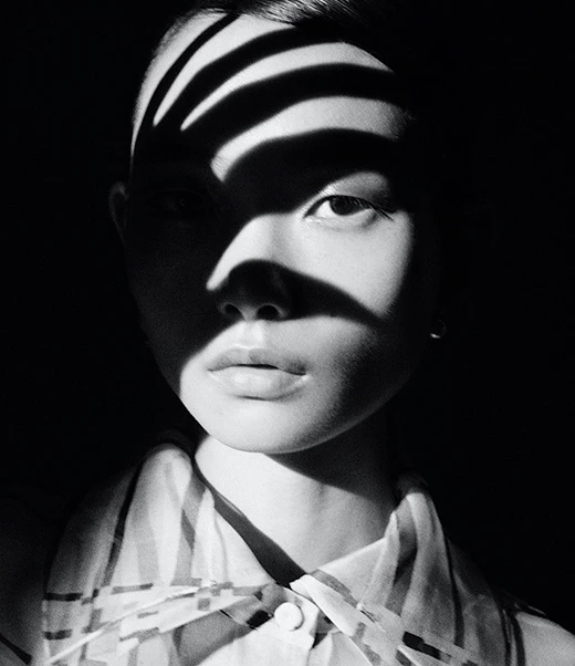 Monochrome portrait of an Asian woman with striped shadows across her face, wearing a striped blouse against a dark background.