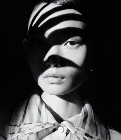 Monochrome portrait of an Asian woman with striped shadows across her face, wearing a striped blouse against a dark background.