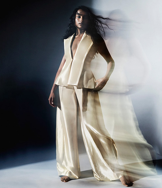 A woman poses in a flowing, off-white two-piece outfit against a smoky dark background, creating a dramatic fashion image with movement.