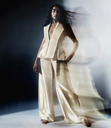 Woman in shiny cream-colored sleeveless top and matching wide-leg trousers posing in studio with motion blur effect.