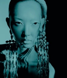 Close-up portrait of a woman wearing oversized jewelled drop earrings under dramatic teal lighting by Paul Scala.