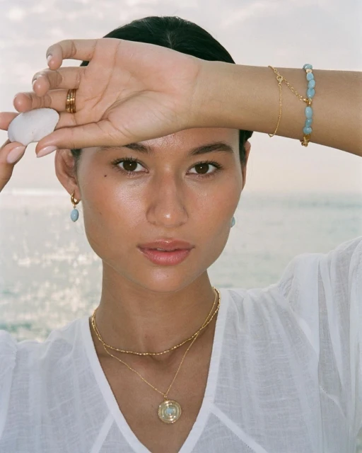 Portrait of a woman with gold jewelry and a white top, posing with a smooth white stone against a soft, out-of-focus ocean backdrop.