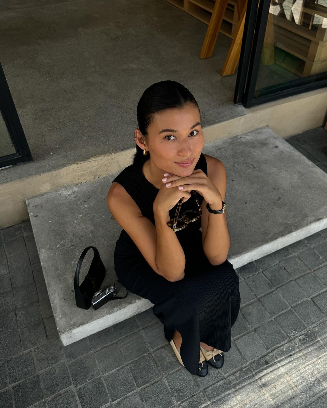 A young woman sits on concrete steps with her hands clasped under her chin, wearing a black dress and flats, with a purse and camera.