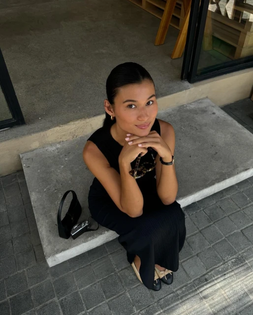 A young woman sits on concrete steps with her hands clasped under her chin, wearing a black dress and flats, with a purse and camera.
