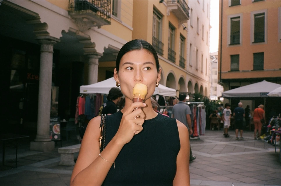 Young woman enjoys an ice cream cone at an outdoor market in a European city, featuring surrounding buildings and vendor stalls.